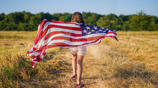 счастливая маленькая девочка-патриотка бежит в поле с американским флагом. - july 4 стоковые фото и изображения