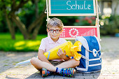 Glückliche kleine Kind Junge mit Brille sitzen durch Schreibtisch und Rucksack oder Tasche. Schulkind mit traditionellen deutschen Schultasche Kegel genannt Schultuete an seinem ersten Tag in die Schule