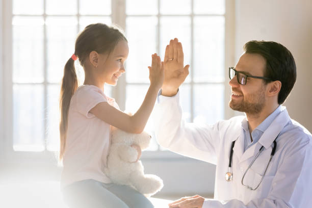 Happy little preschool girl giving high five to male doctor. Side view happy little preschool girl giving high five to male doctor at meeting in hospital. Smiling small patient celebrating successful treatment finish with general practitioner at checkup. general view stock pictures, royalty-free photos & images