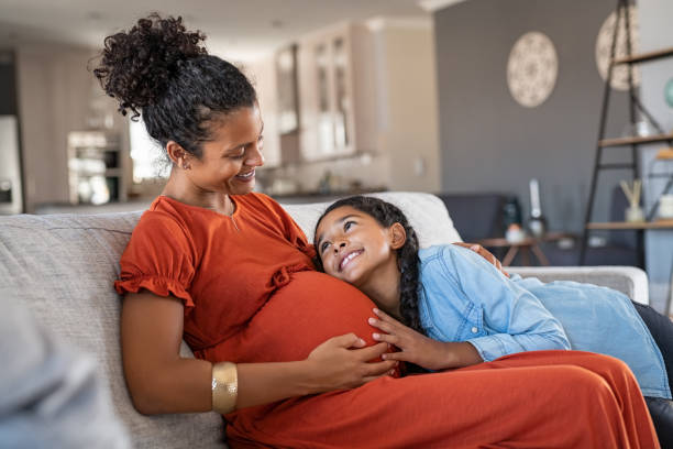 Happy lovely daughter hugging pregnant mother Happy mixed race daughter hugging belly of her expecting mother while relaxing on couch at home. African girl listening to baby movements while embracing pregnant woman. Pregnant black mom and future sister relaxing together on sofa at home. pre pregnancy care stock pictures, royalty-free photos & images