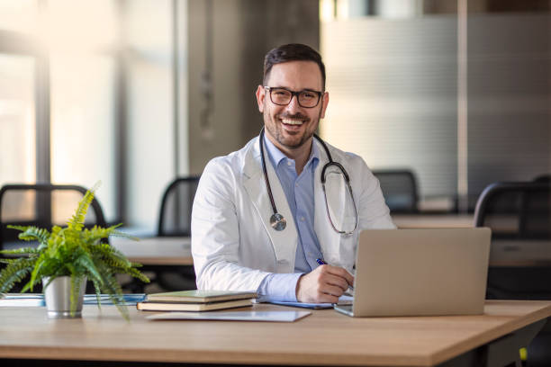 Happy male medical doctor portrait in hospital Happy male medical doctor portrait in hospital. Portrait of a male doctor with laptop sitting at desk in medical office. Portrait of a happy young doctor at medical office desk. doctors office stock pictures, royalty-free photos & images