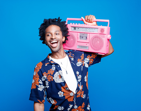 Portrait of excited afro american young man with dreadlocks wearing hawaiian short sleeved shirt holding pink boom box on his shoulder. Studio shot against blue background.