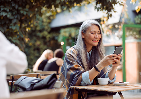 Happy mature Asian woman surfs internet on mobile phone sitting at small table on outdoors cafe terrace on autumn day