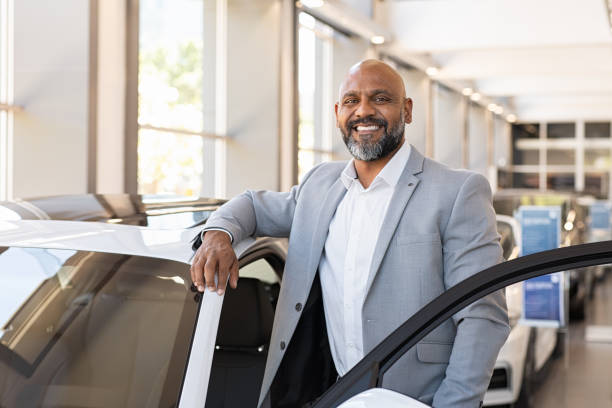 Happy mature black man at car dealership African salesman standing in car showroom near opened door of luxury car. Happy businessman buying his new suv in car salon. Smiling mature man choosing his new car. car salesperson stock pictures, royalty-free photos & images