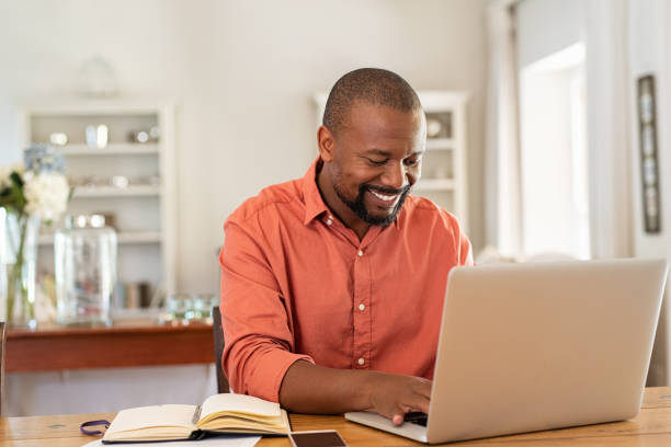 Happy mature black man using laptop Smiling black man using laptop at home in living room. Happy mature businessman send email and working at home. African american freelancer typing on computer with paperworks and documents on table. using laptop stock pictures, royalty-free photos & images