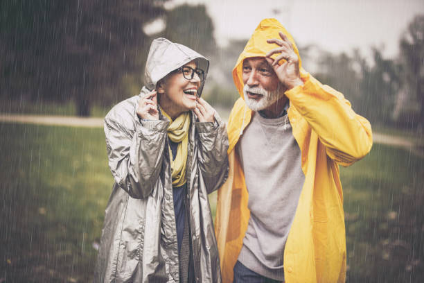 felice coppia matura in impermeabili durante la pioggia nel parco. - autunno energia foto e immagini stock