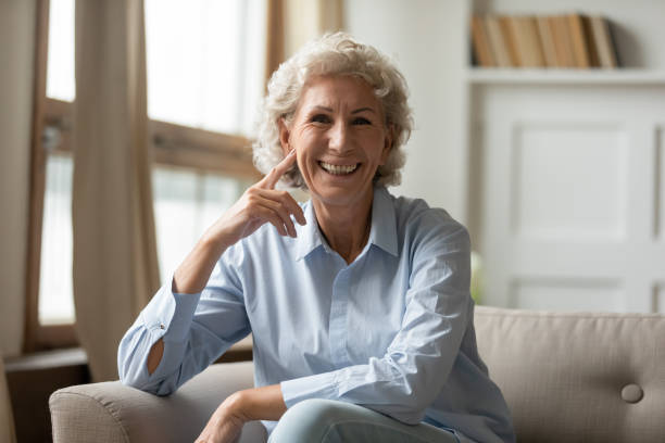 Happy mature granny sitting on couch smiling posing for camera Joyful old woman sit on couch looks at camera, concept of videocall distant chat with family relatives. Granny with toothy wide smile, dentures advertisement dental clinic professional services offer free adult cams stock pictures, royalty-free photos & images