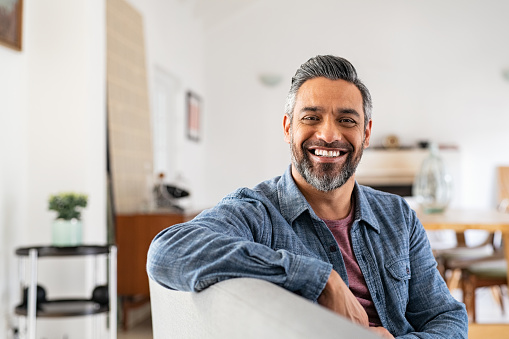 Portrait of handsome mature man relaxing on couch at home while looking at camera. Mixed race man in casual clothing sitting on sofa and smiling with copy space. Successful happy middle eastern guy with beard indoor.