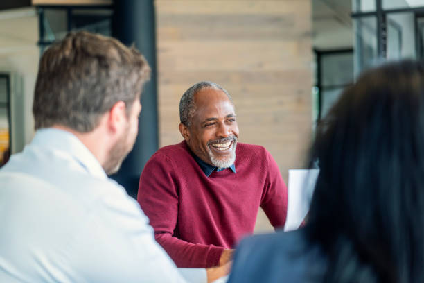 Happy mature male manager sitting with colleagues Smiling mature male manager sitting with colleagues in board room. Multi-ethnic professionals are planning strategy during meeting. They are at new office. business casual stock pictures, royalty-free photos & images