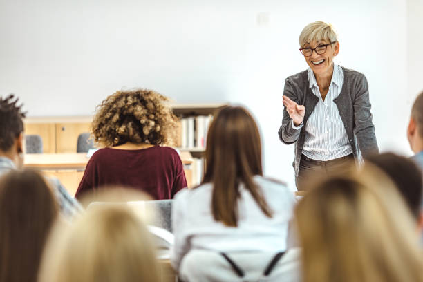 Happy mature professor giving a lecture in front of her students at lecture hall. Happy senior teacher talking to large group of college students in amphitheater. professor stock pictures, royalty-free photos & images