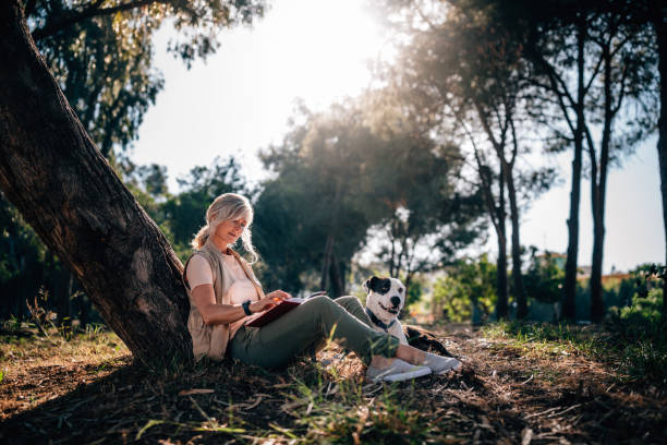 Happy mature woman relaxing in nature with dog and book Fashionable senior woman with pet dog sitting under tree in forest park and reading book wundervisuals stock pictures, royalty-free photos & images