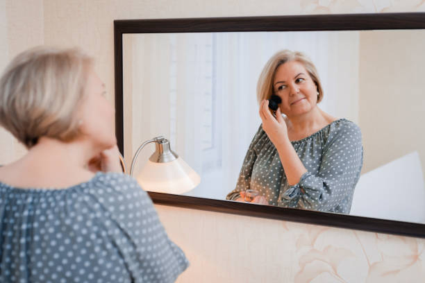happy middle aged elderly woman preens and adjusts her hair in front of the mirror of the dressing table stock photo