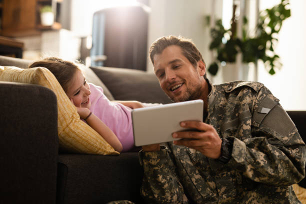 Happy military father and daughter using digital tablet at home. Happy little girl lying on the sofa and watching something on touchpad with her military dad. veterans returning home stock pictures, royalty-free photos & images