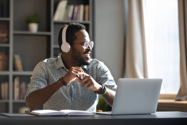 feliz hombre de negocios africano milenario usa auriculares escuchando música en la oficina - auriculares equipo de música fotografías e imágenes de stock