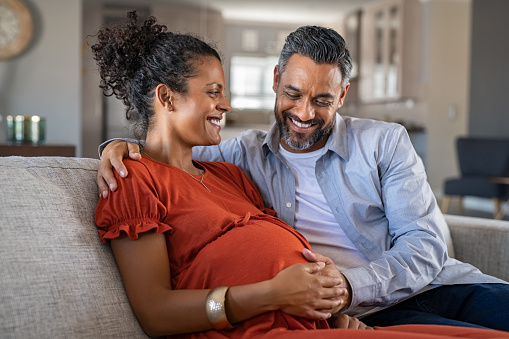 Mid adult couple expecting a baby while husband caresses the belly of his pregnant african woman. Happy father hands on expecting mother's baby bump while embracing her on couch. Lovely indian man touching belly of his girlfriend and feeling baby movement.