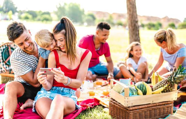 Happy multiethnic families playing with phone at pic nic garden party - Joy and love life style concept with mixed race people having fun together at picnic barbecue before sunset stock photo