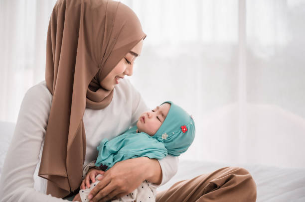 Happy muslim mother holding adorable little baby daughter wearing hijab in her arms on white bed in bedroom. stock photo