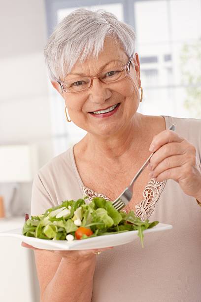 Happy old woman eating fresh green salad, smiling, looking at camera..