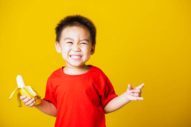 Happy portrait Asian child or kid cute little boy attractive smile wearing red t-shirt playing holds peeled banana for eating, studio shot isolated on yellow background Happy portrait Asian child or kid cute little boy attractive smile wearing red t-shirt playing holds peeled banana for eating, studio shot isolated on yellow background cute thai girl stock pictures, royalty-free photos & images