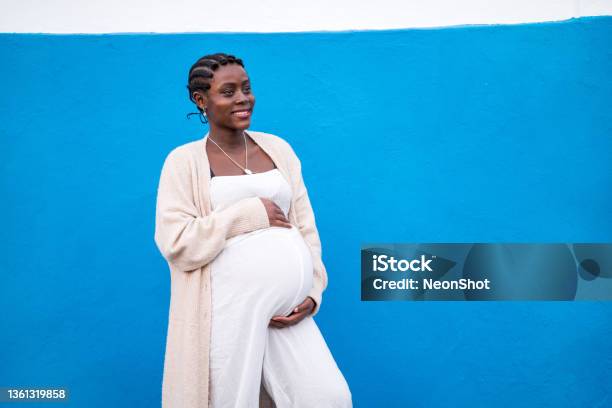 Happy pregnant african woman holding her belly in hands, smiling, posing on the blue wall outdoor. Real people lifestyle and emotions. Maternity.