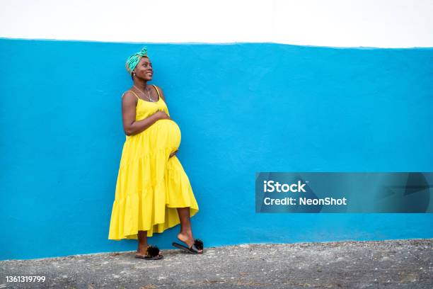 Happy pregnant african woman in yellow dress holding her belly in hands, smiling, posing on the blue wall outdoor. Real people lifestyle and emotions.