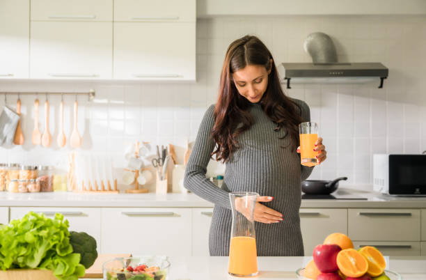 Happy pregnant woman in maternity dress standing holding a glass of orange juice and looking down at belly in kitchen at home. stock photo