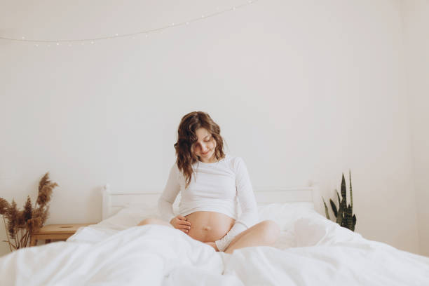 Happy pregnant woman in white holding belly bump and relaxing on white bed at home. Stylish pregnant mom waiting for baby. Motherhood and fertility concept. Maternity time stock photo