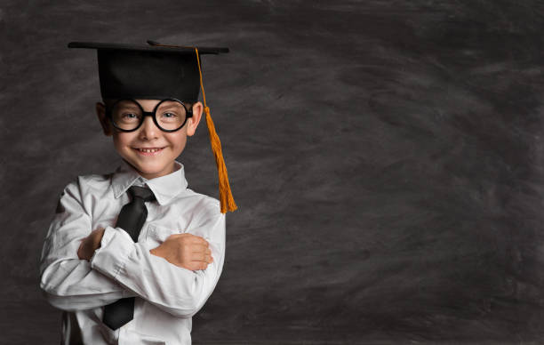 Happy Preschooler Boy in Eyeglasses and Graduation Hat over Blackboard with Copy Space. Happy Little Kid like Student in Smart Casual White Shirt Black Tie. Clever Child in Master Cap stock photo