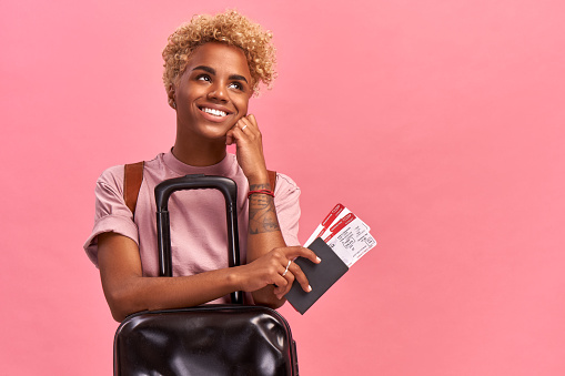 Happy pretty Afro female tourist with Luggage holds passport and plane tickets, rejoices having tour abroad with boyfriend, has special offer from travel company, on pink background.