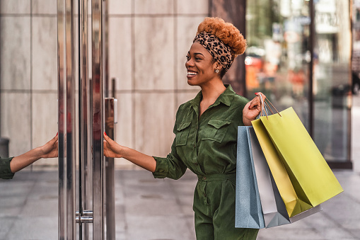 Stylish Afro American woman holding shopping bags and opening the door of a shopping mall in the city while smiling. Lifestyle concept