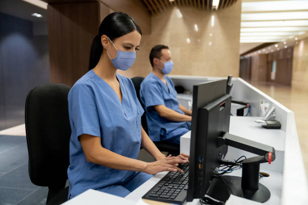 Happy receptionist working at a hospital wearing a facemask Happy receptionist working at a hospital wearing a facemask during the COVID-19 pandemic - healthcare and medicine concepts hotel reception photos stock pictures, royalty-free photos & images