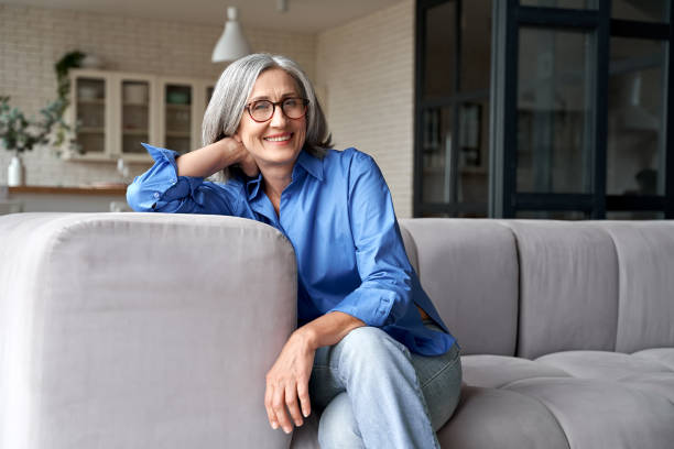 Happy relaxed mature old adult woman wearing glasses resting sitting on couch at home. Smiling mid age grey-haired elegant senior lady relaxing on comfortable sofa looking at camera. Portrait Happy relaxed mature old adult woman wearing glasses resting sitting on couch at home. Smiling mid age grey-haired elegant senior lady relaxing on comfortable sofa looking at camera. Portrait 50 59 years photos stock pictures, royalty-free photos & images