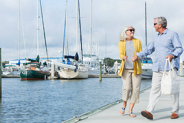 Happy senior couple walking along harbor holding hands A happy, senior couple walking together holding hands along a boat dock, enjoying the water and view of the luxury sailboats on a sunny summer day. They are smiling, face to face, enjoying retirement. marina stock pictures, royalty-free photos & images