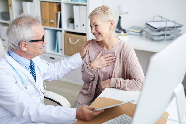 Happy senior female patient with short hair sitting at table in doctors office and holding hand on chest while thanking doctor for good treatment and support Happy senior patient thanking doctor russian mature women stock pictures, royalty-free photos & images
