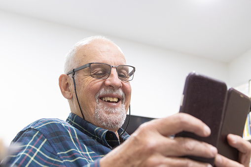 Happy senior man in home office using mobile phone and smiling: relaxed man has white hair and beard, wearing eye glasses with toothy grin. Interaction, communication. Phone ID and camera removed.
