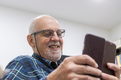 Happy senior man in home office using mobile phone and smiling: relaxed man has white hair and beard, wearing eye glasses with toothy grin. Interaction, communication. Phone ID and camera removed.