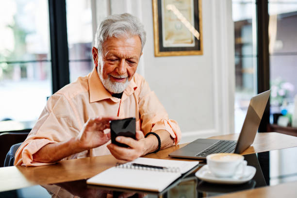 A happy senior student is sitting in a cafe and using his phone to check up on updates related to school. On the table are a laptop and notebook. stock photo