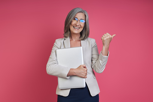 Happy senior woman in formalwear carrying laptop and pointing away against pink background