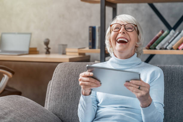 glückliche seniorin schaut und lacht über ihr digitales tablet auf dem sofa - lachen stock-fotos und bilder