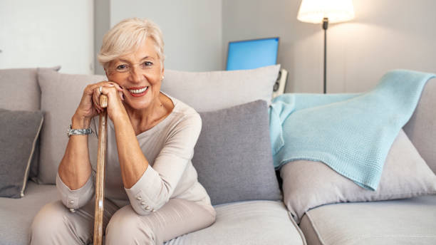Happy senior woman with walking stick seating at home. Smiling grandmother sitting on couch. Portrait of a beautiful smiling senior woman with walking cane on light background at home. Old woman sitting with her hands on a cane senior women stock pictures, royalty-free photos & images