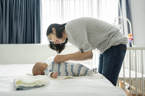 Happy Smiling Asian Young mother playing and dressing her little baby son on bed. stock photo