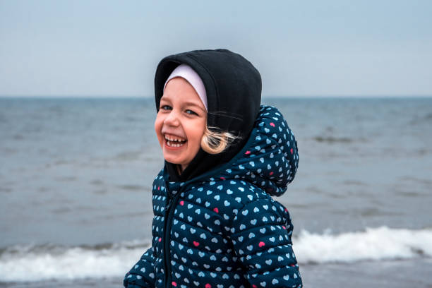 Happy smiling beautiful child girl in warm clothes walking on the beach in cold autumn day with sea on background in blue colours as happy childhood lifestyle stock photo