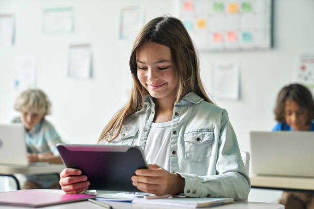 Happy smiling schoolgirl reading task from tablet computer device in classroom. stock photo