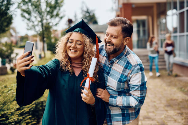 Happy student taking selfie with her father on her graduation day. Happy university graduate and her father having fun while taking selfie with smart phone. online phd programs stock pictures, royalty-free photos & images
