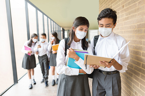 Happy students wearing facemasks at the school while talking in the hall sharing notes - COVID-19 pandemic concepts