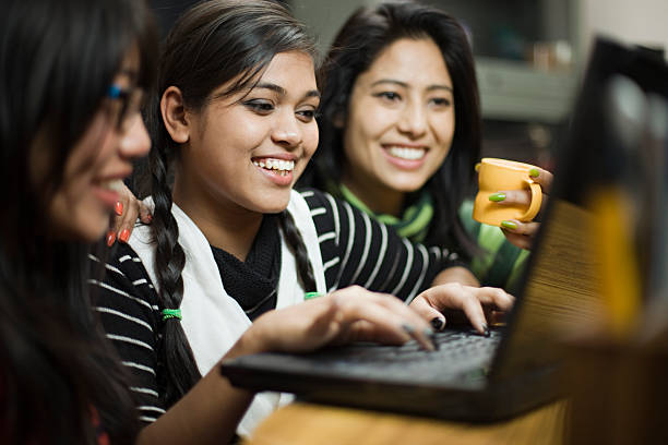 Happy teenage Asian girls of different ethnicity using laptop together. Indoor image of a group of late teen girls from different ethnicity using laptop together. Selective focus is on Indian girl sitting in the middle and typing on the keyboard while looking at laptop screen. Group of people, waist up, horizontal composition with selective focus. asian girl stock pictures, royalty-free photos & images
