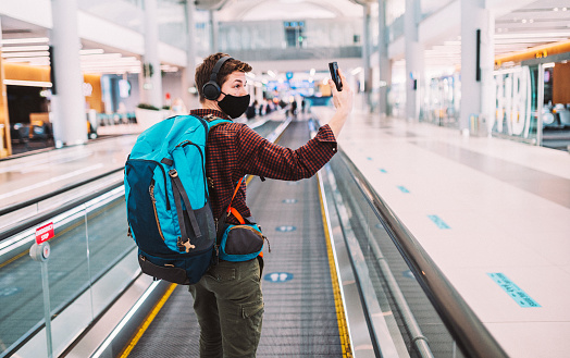 Rear view of a young man traveler, in a protective face mask, doing selfie, video chatting on a smartphone, while he is standing on an elevated walkway at the airport with a social distancing sign on it.\nTraveling during the Coronavirus pandemic