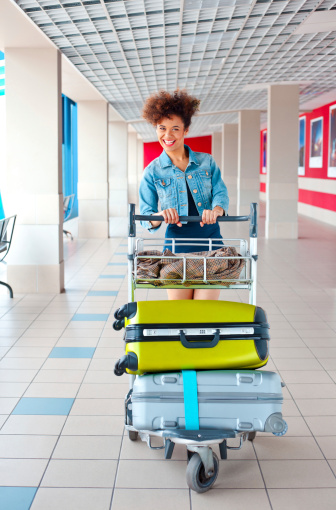 Portrait of teenaged girl pushing an luggage trolley with her suitcases at the airport and laughing at the camera.