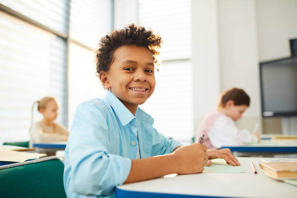 Happy Twelve-Year-Old Student Smiling Horizontal low angle medium close up portrait of happy mixed-race boy with kinky hair sitting at school desk looking at camera smiling, copy space middle school teacher stock pictures, royalty-free photos & images