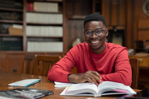 Happy university student in library Portrait of university student doing homework in school library and smiling. Happy high school student looking at camera while studying for exam. African american clever guy with open book sitting at desk with copy space. college students stock pictures, royalty-free photos & images